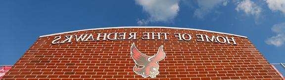 A brick wall that says "Home of the Redhawks" and has the Roberts Redhawk logo. The blue sky is visible above with some white clouds.